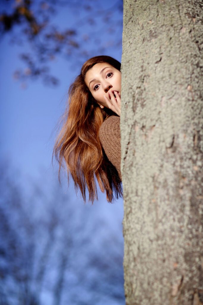 Junge Frau schaut überrascht hinter einem Baum hervor